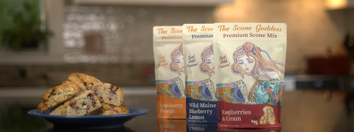 Three packages of 'The Scone Goddess' scone mix with a plate of scones on a kitchen counter.